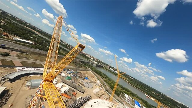 Yellow tower crane lifts freights near concrete stadium building at construction site aerial camera rotates and lowers