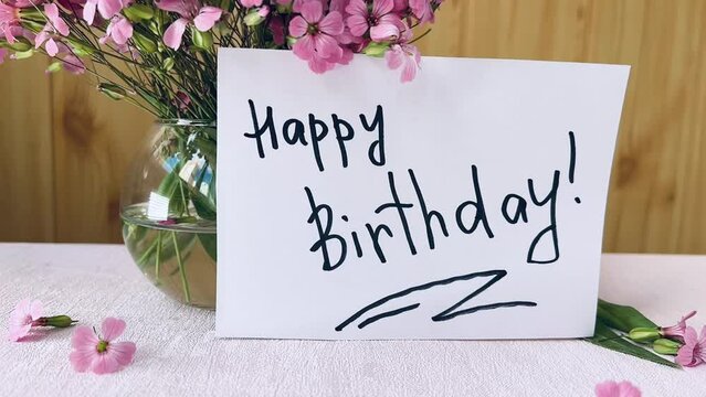 Happy birthday card with greeting words and bouquet of pink flowers on wooden background in slow motion