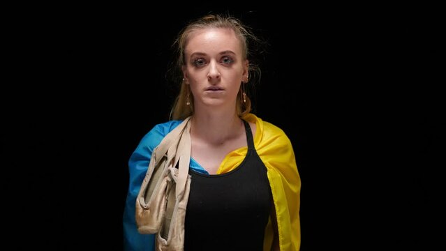 Sad young woman in Ukrainian flag with pointies hanging on shoulder looking at camera. Portrait of frustrated lady posing at black background with hopeless facial expression