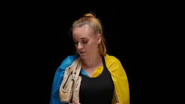 Slim graceful ballerina in Ukrainian flag with pointies on shoulder walking from darkness looking at camera. Portrait of brave confident woman posing at black background. Art and war