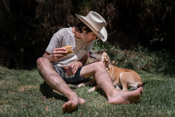 A Mexican young man wearing a hat is eating a taco and playing with his dog sitting on the grass.