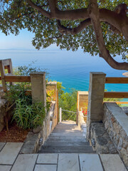 Walking through a beautiful restaurant terrace on the shores of the Mediterranean Sea in Greece. Seaside terrace with vegetation and flowers ending in stairs that descend into the clear turquoise sea.