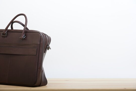 An Elegant Business Briefcase Made Of Old Craft Dark Brown Leather Stands Next To The White Wall As A Background On The Smooth Surface Of Light Wooden Table. No People Studio Photography.