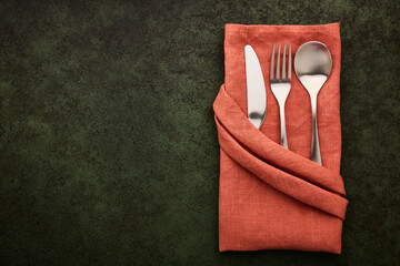 Terracotta-colored linen napkin folded in the shape of a pocket with cutlery inside, dark green background with copy space - 515037432
