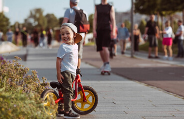 Funny little boy riding a bicycle (running bike) outdoors in the city. A happy child walks in the park. The kid is wearing sweatpants, a T-shirt and sneakers.