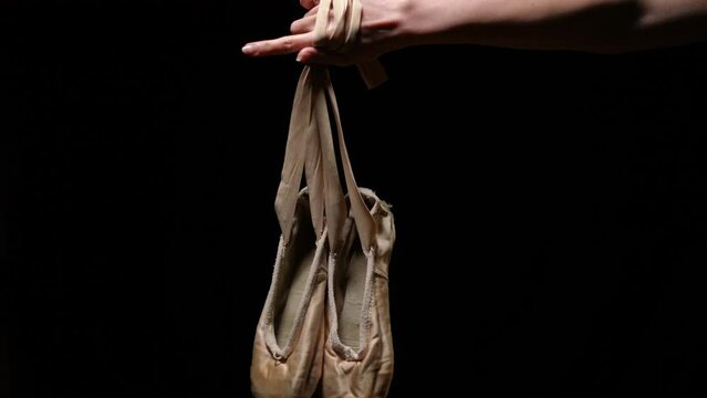 Ballet shoes hanging on slim female hand at black background close-up. Pointies on palm of unrecognizable confident ballet dancer. Choreography and art concept