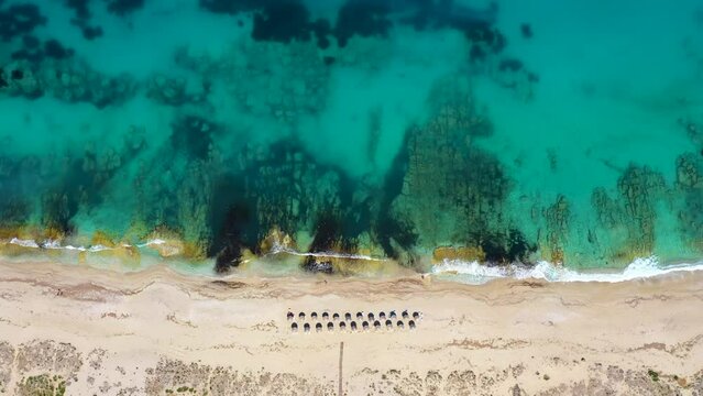 Umbrellas lined up on the sandy beach by the turquoise sea. Top view of a transparent blue sea with beautiful waves on the beach. Summer vibes concept. Exotic beach from above