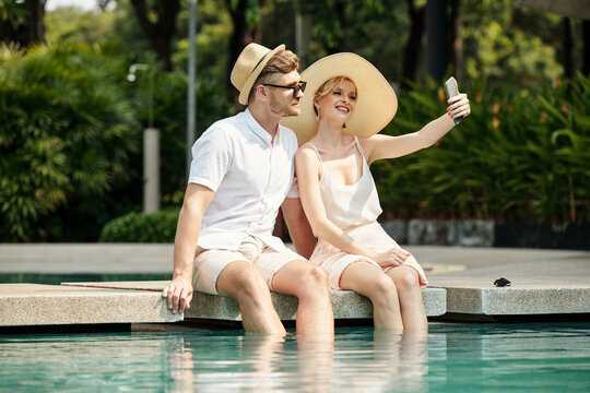 Young adult Caucasian couple enjoying spending time together at hotel territory sitting at pool with legs in water taking selfie on smartphone