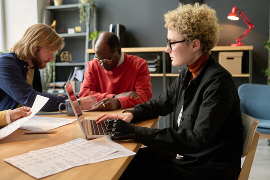 Young Businesswoman With Prosthetic Arm Using Laptop Sitting At Table With Her Colleagues During Business Meeting At Office