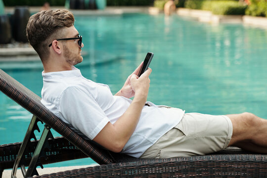 Side view of young adult Caucasian man relaxing on deck chair at poolside surfing Internet on smartphone on summer day