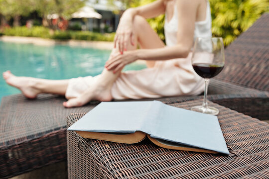 Selective Focus Shot On Table With Book And Glass Of Wine On It, Unrecognizable Woman Sitting Relaxed On Deck Chair At Poolside On Background