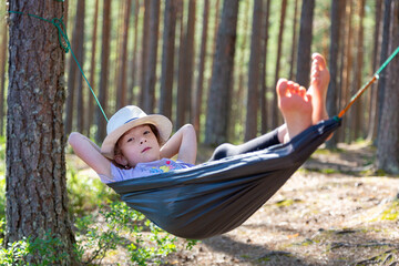 A child in a hat and barefoot sits in a hammock in pine wood, looks at the lake. Country style