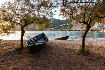 Naklejka na ściany i meble Two small boats lying on the sea shore under low trees on a rocky beach. In the background the sea and the promontory of the mainland.