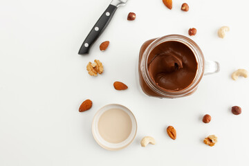 A jar of chocolate paste, a lid, a knife and various types of nuts. Cashew, almond, walnut, hazelnut, isolated on white background.