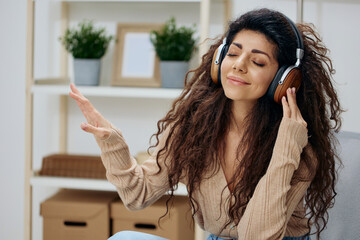 Relaxed calm pleasant tanned curly Latin lady with headphones moves to cool music sit on chair in...