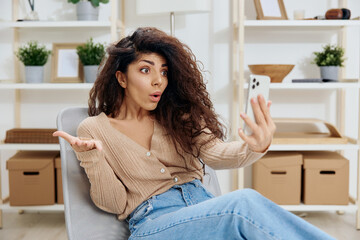 Confused unhappy irritated shocked tanned curly Latin lady have video call hold trendy smartphone sit on chair in home modern interior look at screen. Copy space Mockup Banner. Concept shock info