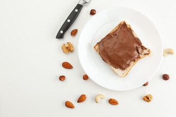Bread with chocolate paste on a plate. Knife and various types of nuts. Cashew, almond, walnut, hazelnut isolated on white background.