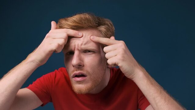 Face close up of a young ginger man looking at his skin problems examining his face over dark blue background.