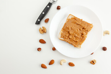 Bread with peanut butter on a plate. Knife and various types of nuts. Cashew, almond, walnut, hazelnut isolated on white background.