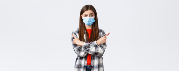 Fototapeta na wymiar Coronavirus outbreak, leisure on quarantine, social distancing and emotions concept. Indecisive and clueless pretty girl in medical mask ask your opinion, advice as pointing sideways left and right