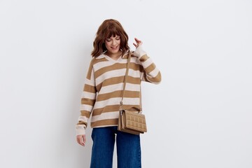 a close photo of a beautiful, stylish woman in a striped sweater and blue jeans posing relaxed standing on a light background with a bag worn over her shoulder looking to the side