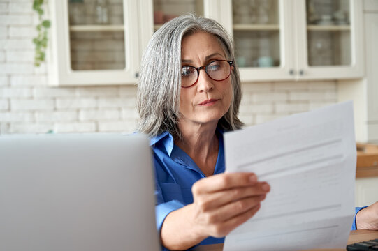 Serious worried mature woman reading paper bill pay online at home. Old lady holding bank letter managing account finances, calculating taxes, planning loan debt pension payment sit at kitchen table.