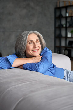 Smiling relaxed mature old woman resting dreaming sitting on couch at home. Happy mid aged woman relaxing. Peaceful serene grey-haired lady feeling peace of mind, no stress on sofa, vertical view.