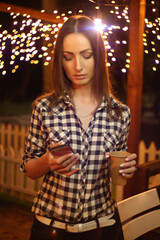 beautiful fashionable girl in a plaid shirt is drinking coffee and looking at the phone against the background of lanterns