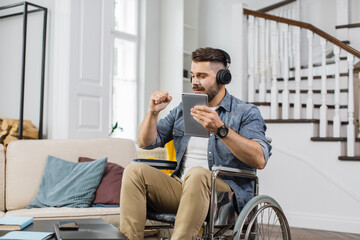 Caucasian man in wheelchair wearing headphones and slightly moving under dancing playlist on tablet app. Young male with special needs enjoying favorite songs in modern living room.