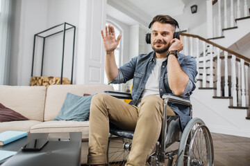 Fototapeta na wymiar Disabled man in modern headphones waving hand while looking at camera with smile. Pleasant guy sitting in wheelchair and listening good music at home.