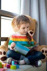 A cute girl of 6 years old sits on the couch by the window with a large toy bear and reads a book....