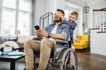 Happy son unexpectedly closing father's eyes from back in modern living room. School boy surprising disabled dad in wheelchair with smartphone in hands.