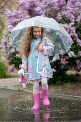 A sweet girl of five years with a transparent umbrella against the background of blooming lilacs enjoys spring and has a fun. Pastel colors. Spring. Childhood. Beauty.