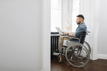 Fototapeta na wymiar Male freelancer with disability working on wireless laptop in bright room at home. Bearded man wearing casual clothes sitting in wheelchair near big window.
