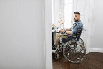 Fototapeta na wymiar Focused bearded man who using wheelchair holding laptop on knees and looking at camera. Hardworking male freelancer with disability working remotely from home.