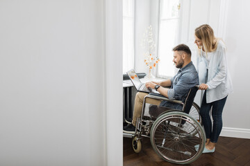 Fototapeta na wymiar Disabled caucasian man sitting in wheelchair and working on modern laptop while his charming wife standing behind. Family support, people and love.