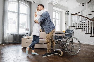 Caucasian female rehabilitation specialist hugging handsome man after successful physical therapy in living room. Concept of medical care, people, support and disability.