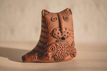Clay figurine of a cat with a fish on a light background close-up. Crafts made of clay. A cat holds...
