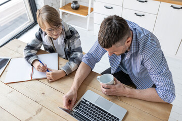 Father helping his teenage son with homework while working from home in the kitchen. Concept of parenthood, fatherhood, spending quality time together. Using technology, gadgets, devices for learning