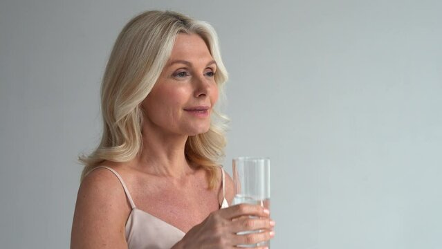 Healthy thirsty fit mid aged 50s woman holding glass drinking water. Happy mature lady hydrating body with clean filtered fresh mineral water for skin care, beauty, diet nutrition hydration.