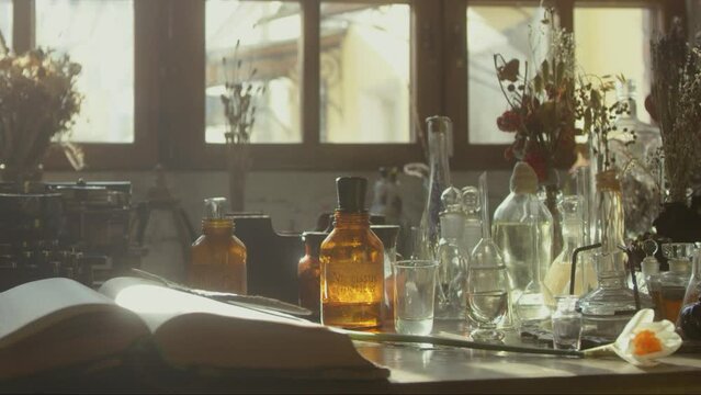 Close-up of a perfumer's workplace. Vintage cinematic concept. Sunny day in the working workshop. Lots of ingredients, a book, glass flasks on the table. Depth of field. 4K video