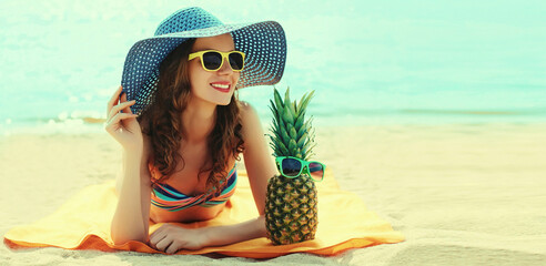 Summer portrait of happy smiling young woman lying on the beach with funny pineapple wearing straw...