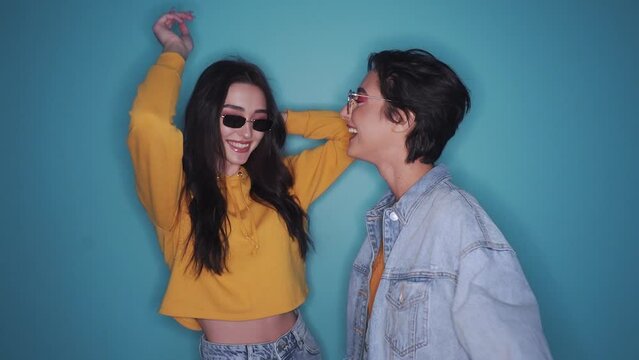 Two happy stylish cool young women wearing sunglasses having fun dancing, laughing, at party on blue background in slow motion.