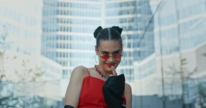 Portrait of stylish gorgeous cheeky woman, seductive happy female wearing cool red eyeglasses smiles to camera and flirts standing in modern business center overlooking high rise glass skyscrapers