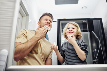 Low angle portrait of smiling father and son shaving together and looking in mirror during morning routine