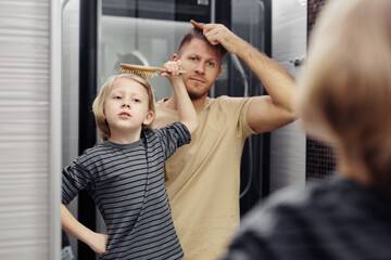 Portrait of loving father and son bushing hair together and looking in bathroom mirror, morning...