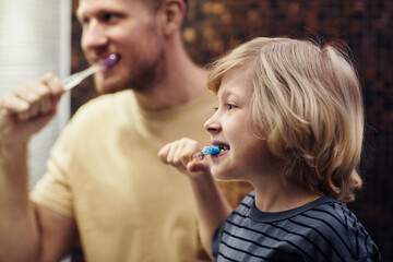 Side view portrait of cute blonde boy brushing teeth with father in background, both looking in mirror