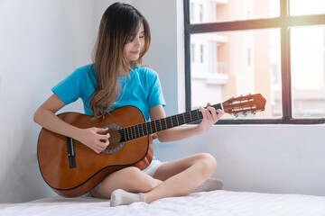 Cheerful asian woman casual playing acoustic guitar sitting on white bed beside window at...
