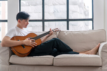 Happy asian handsome man playing acoustic guitar sitting on sofa relax beside window at apartment....