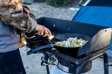 Woman Cooking potatoes on outdoor camping propane stone using a knife as a utensil with the sun...